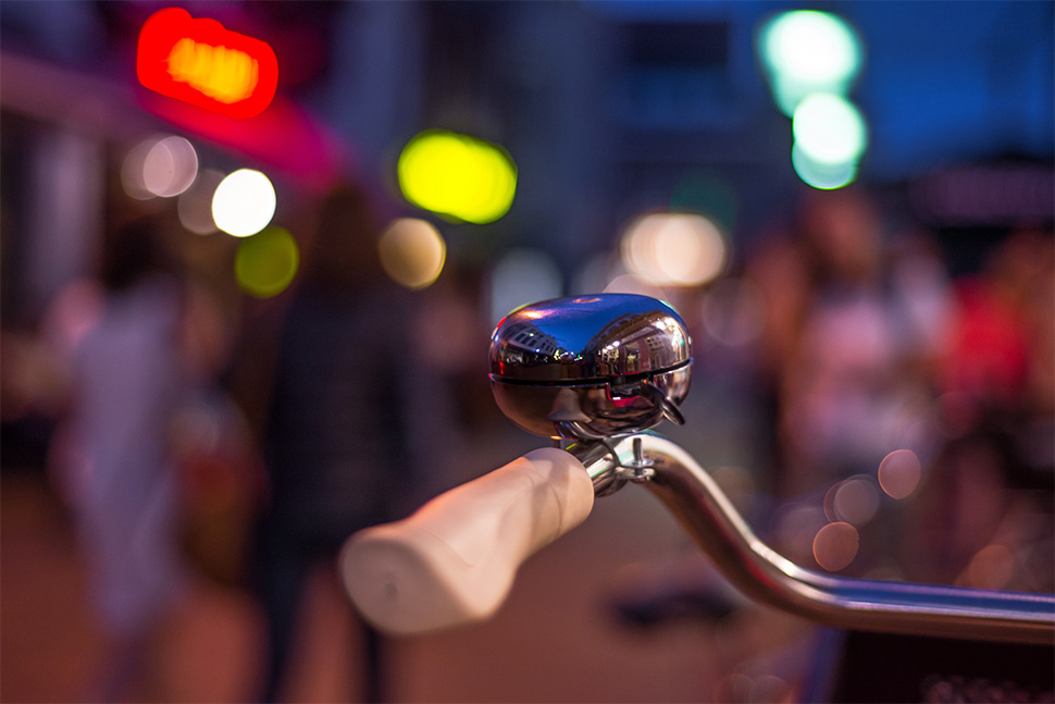 Fahrradlenker mit großer Klingel, im Hintergrund Lichter der Straße